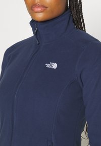 Marineblaue Fleecejacke mit hohem Kragen, Frontreißverschluss und gesticktem Logo auf der Brust. Glatte Textur und taillierte Passform.