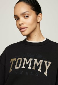 Sudadera negra con la frase "TOMMY JEANS" en letras brillantes doradas y azul marino, con un cuello redondo y un interior del cuello de color claro.