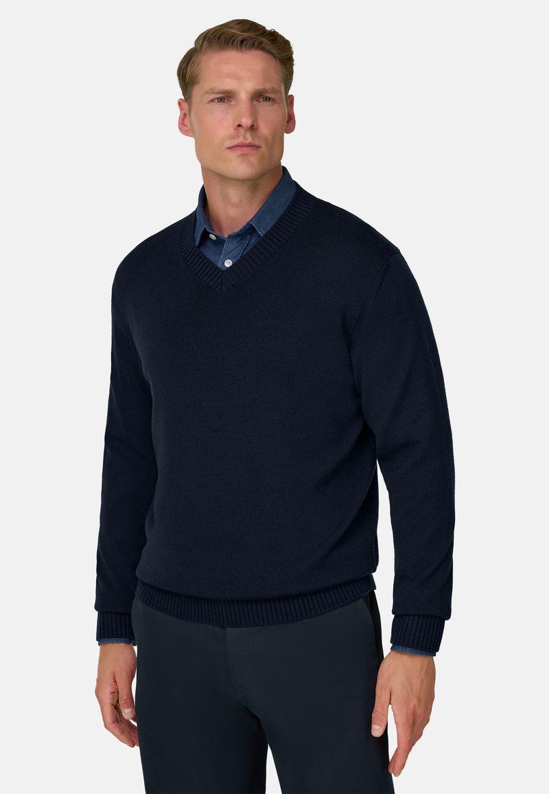 Homme portant un pull bleu marine à col en V par-dessus une chemise bleue boutonnée, debout devant un fond clair uni.
