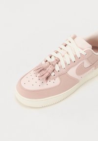 Zapatilla rosa claro con puntera perforada, cordones blancos y acentos de borlas de tela. Materiales lisos y texturizados con paneles en contraste.