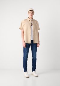 Camicia beige a maniche corte con bottone davanti, indossata sopra una maglietta bianca, abbinata a jeans blu scuro e sneakers crema, completata da un berretto lavorato a maglia.