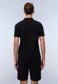 Svart polo skjorte med en fast tekstur, korte ermer og en subtil rød logodetalj ved bakre krage. Kombinert med svarte shorts.
