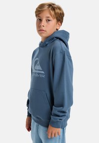 Blue hoodie with a front kangaroo pocket, drawstring hood, and a large white logo on the chest, made of soft fabric, fitted sleeves.