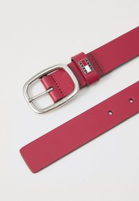 Red leather belt with a silver rectangular buckle. Features a logo plate on the buckle with "TOMMY JEANS" and multiple adjustment holes on the strap.