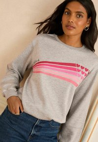 Sudadera gris de manga larga, con un diseño de corazones a rayas rosas y el texto "c'est la vie". Llevada con jeans azul oscuro y un anillo.
