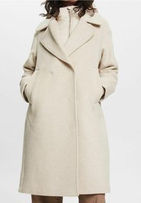 Abrigo de mezcla de lana beige con cuello de solapa, diseño de doble botonadura, bolsillos laterales y textura suave, superpuesto a una chaqueta interior ligeramente acolchada.