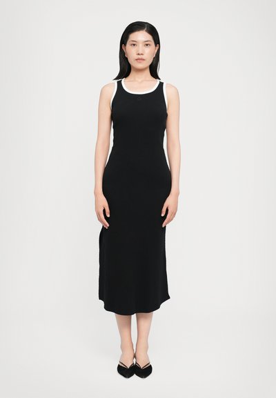 Robe sans manches noire à côtes avec des bords blancs, silhouette ajustée, longueur genou, fentes latérales, associée à des chaussures à talons pointus noires.
