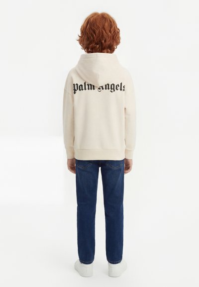 Laps, kellel on lokkis punased juuksed, kannab kreemikat kapuutsiga pusa, millel on taga kirjas "Palm Angels", siniseid teksapükse ja valgeid tossusid, seiseb seljaga vaataja poole.
