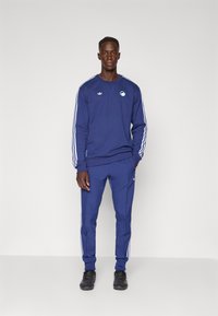 Tumši zils sporta krekls un atbilstošas sporta bikses ar baltām trīs svītru uzrakstēm. Uz krūtīm ir uzšūts logotips, kā arī sānu kabatas. Izgatavots no mīksta materiāla.