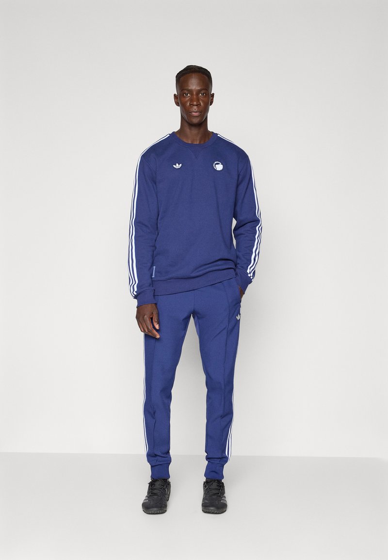 Marineblauer Trainingsanzug mit weißen Streifen, bestehend aus einem Rundhalsausschnitt-Pullover und schmal zulaufenden Hosen. Enthält Logo-Akzente auf der Brust und der Hüfte.