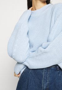 Vaaleansininen neulepusero, jossa on ribbikuvio, ylisuuri malli ja leveät hihat. Yhdistetty korkeavyötäröisiin farkkuihin.