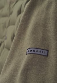 Olive green fabric with a smooth texture features a dark blue rubber label embossed with the text "bugatti." Background includes quilted design.