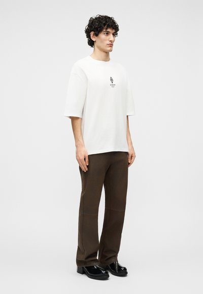 Άνδρας με κατσαρά μαλλιά, που φοράει άσπρο, φαρδύ T-shirt, σκούρο καφέ παντελόνι και μαύρα δερμάτινα παπούτσια, στέκεται μπροστά σε απλό φόντο.