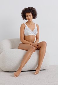 Bijeli čipkani bralet s cvjetnim uzorkom, koji ima podesive naramenice i V-izrez. Model sjedi na mekanoj, zaobljenoj stolici s golim nogama.