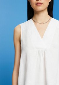 Weiße, ärmellose Bluse mit V-Ausschnitt, plissiertem Detail vorne, aus strukturiertem Stoff gefertigt, kombiniert mit einer silbernen Kettenhalskette.