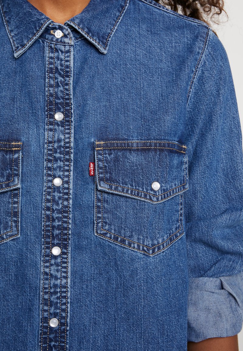 La chemise en denim présente une couleur bleue avec une texture légèrement délavée, deux poches poitrine et des boutons pression en forme de perles sur le devant.