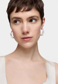 Pendientes aros claros con un acabado brillante, mostrando una forma redonda. El fondo destaca una blusa de tela clara y texturizada.