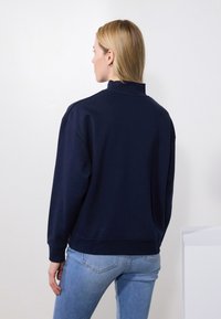 Sudadera de color azul marino con un cuello alto, corte holgado y puños de canalé. Textura suave y diseño sencillo, combinada con jeans de color azul claro.