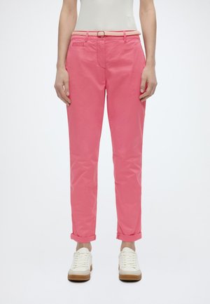 Femme portant un pantalon rose retroussé avec une ceinture tressée et des baskets blanches, debout devant un fond uni.