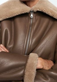 Giacca in pelle marrone con colletto in shearling beige, dotata di chiusura frontale con zip e maniche ampie, che mette in risalto una texture morbida e un design strutturato.