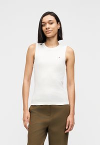 Λευκό αμάνικο τοπ με στρογγυλή λαιμόκοψη, απαλό ύφασμα και μικρή λεπτομέρεια με λογότυπο στο στήθος, συνδυασμένο με καφέ παντελόνι ραμμένο κατά παραγγελία.