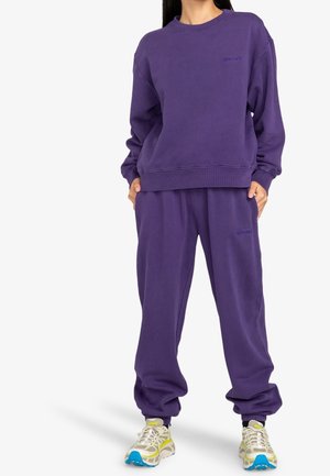 Person wearing loose-fitting purple sweatshirt and matching sweatpants with white and yellow sneakers, standing with hands in pockets.