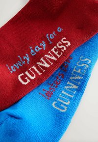 Sarkanas un zilas zeķes ar adītu tekstu: baltā krāsā "lovely day for a GUINNESS" un krēmkrāsā "tastes like GUINNESS", ar gludu tekstūru.