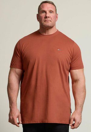 Muskularny mężczyzna stoi na jednolitym tle, ubrany w jednolitą, rdzawego koloru koszulkę z krótkim rękawem i czarne spodnie.