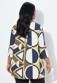 Donna con lunghi capelli neri che indossa una giacca larga con motivo geometrico nei colori blu navy, beige, marrone e senape, rivolta verso un muro bianco.