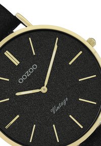 Cadran de montre texturé noir avec des marquages et des aiguilles dorés. Boîtier en métal doré et bracelet noir. Présente le texte 'oozoo' et 'Vintage' en argent.