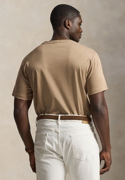 Hátulról látható férfi, akinek bézs rövid ujjú ingje fehér nadrágba van beletűrve, barna övvel, bézs háttér előtt.