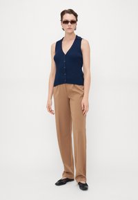 Marineblauw geribd mouwloos shirt met een V-hals en kraag, gecombineerd met lichtbruine broeken. Het model draagt zwarte platte schoenen en een zonnebril.