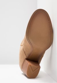 Stivale alla caviglia in suede beige con suola in gomma testurizzata. Il tacco presenta un design in legno e un accento angolato per garantire stabilità. Marchio ALDO visibile.