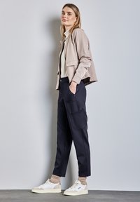 Veste courte beige clair, haut blanc ajusté, pantalon cargo bleu marine avec poches, et baskets blanches avec accents beiges. Texture lisse dans l'ensemble.