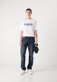 White short-sleeve t-shirt with blue graphic logo, paired with dark blue jeans and white sneakers. Model holds a small black bag.