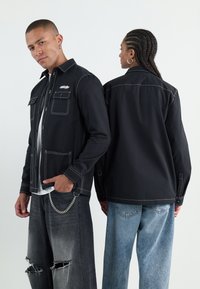 Schwarze Button-up-Jacke mit kontrastierendem weißen Nähten, zwei Fronttaschen, getragen mit verwaschenen grauen Jeans und einem weißen Hemd.