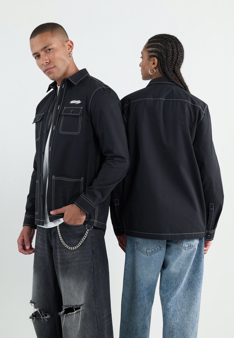 Schwarze Button-up-Jacke mit kontrastierendem weißen Nähten, zwei Fronttaschen, getragen mit verwaschenen grauen Jeans und einem weißen Hemd.