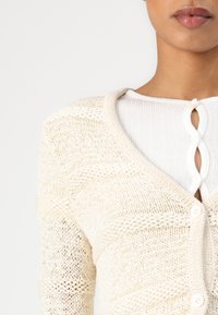 Knit cardigan in cream, featuring a v-neckline, textured patterns, and button closure, layered over a ribbed white top with a keyhole detail.
