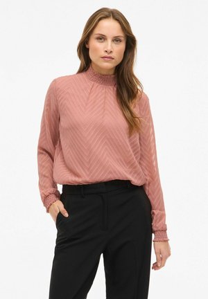 Rosa teksturert bluse med chevronmønster, høy rynket hals og rynkede mansjetter, kombinert med svarte skreddersydde bukser.
