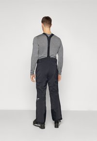 Pantalons de ski noirs imperméables avec bretelles réglables, ceinture élastique et zips latéraux. Portés par-dessus une chemise à manches longues grise.