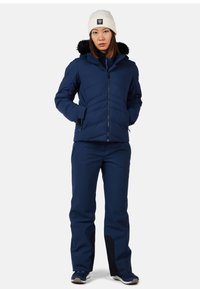 Giacca imbottita blu marina con cappuccio bordato in pelliccia, abbinata a pantaloni blu marina coordinati. La giacca presenta una chiusura con zip anteriore e tasche laterali.