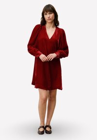 Rode fluwelen jurk met een V-hals, lange pofmouwen en een losse pasvorm, die boven de knieën eindigt. Gecombineerd met bruine platte schoenen.