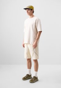 Beige short-sleeve shirt and light shorts, paired with olive sneakers and a two-tone cap. The outfit features a relaxed fit and subtle accents.
