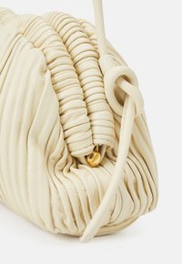 Krémová kožená kabelka s skládaným designem, zaobleným tvarem a uzlovým popruhem. Obsahuje malý detail zlatého zapínání. Hladký povrch.