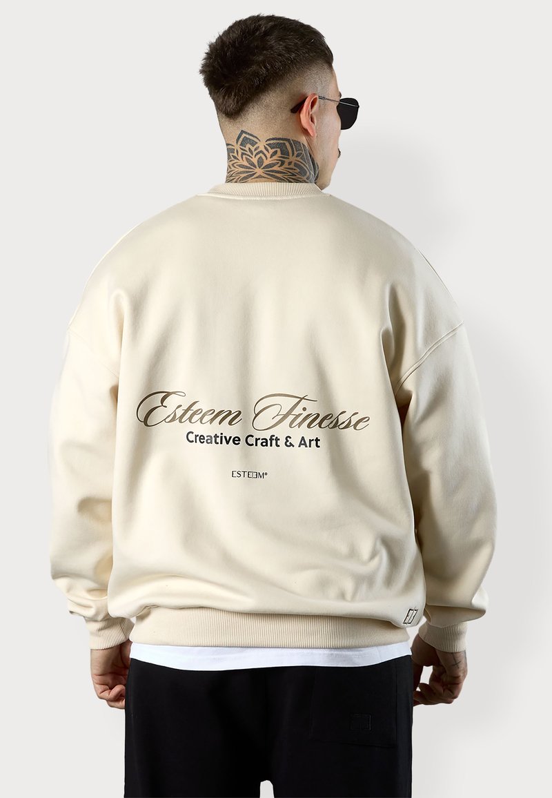 Sweatshirt surdimensionné couleur crème avec un col rond côtelé. Présente un texte imprimé au dos : "Esteem Finesse Creative Craft & Art."