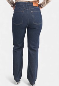 Jeans in denim scuro con taglio a gamba dritta, con una toppa del marchio in pelle sulla cintura e cuciture arancioni. Sono visibili due tasche posteriori.