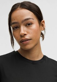 Zwarte t-shirt met een ronde halslijn, gladde textuur en minimale stikdetails, gedragen door een persoon met halflang haar.