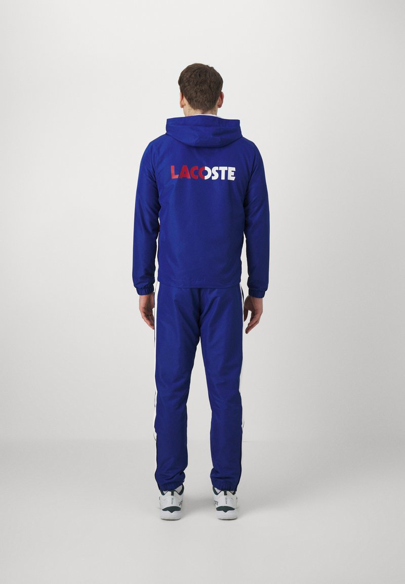 Tuta blu con cappuccio, caratterizzata da un grande logo "LACOSTE" in rosso e bianco sul retro. Strisce laterali bianche sui pantaloni. Vestibilità casual.