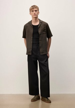 Young man stands against plain wall wearing dark short-sleeve shirt, black tank top, wide-leg black pants, and olive green sneakers.