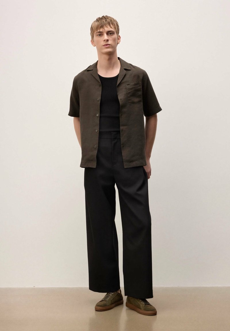 Jeune homme appuyé contre un mur uni, portant une chemise sombre à manches courtes, un débardeur noir, un pantalon noir à jambes larges et des baskets vert olive.